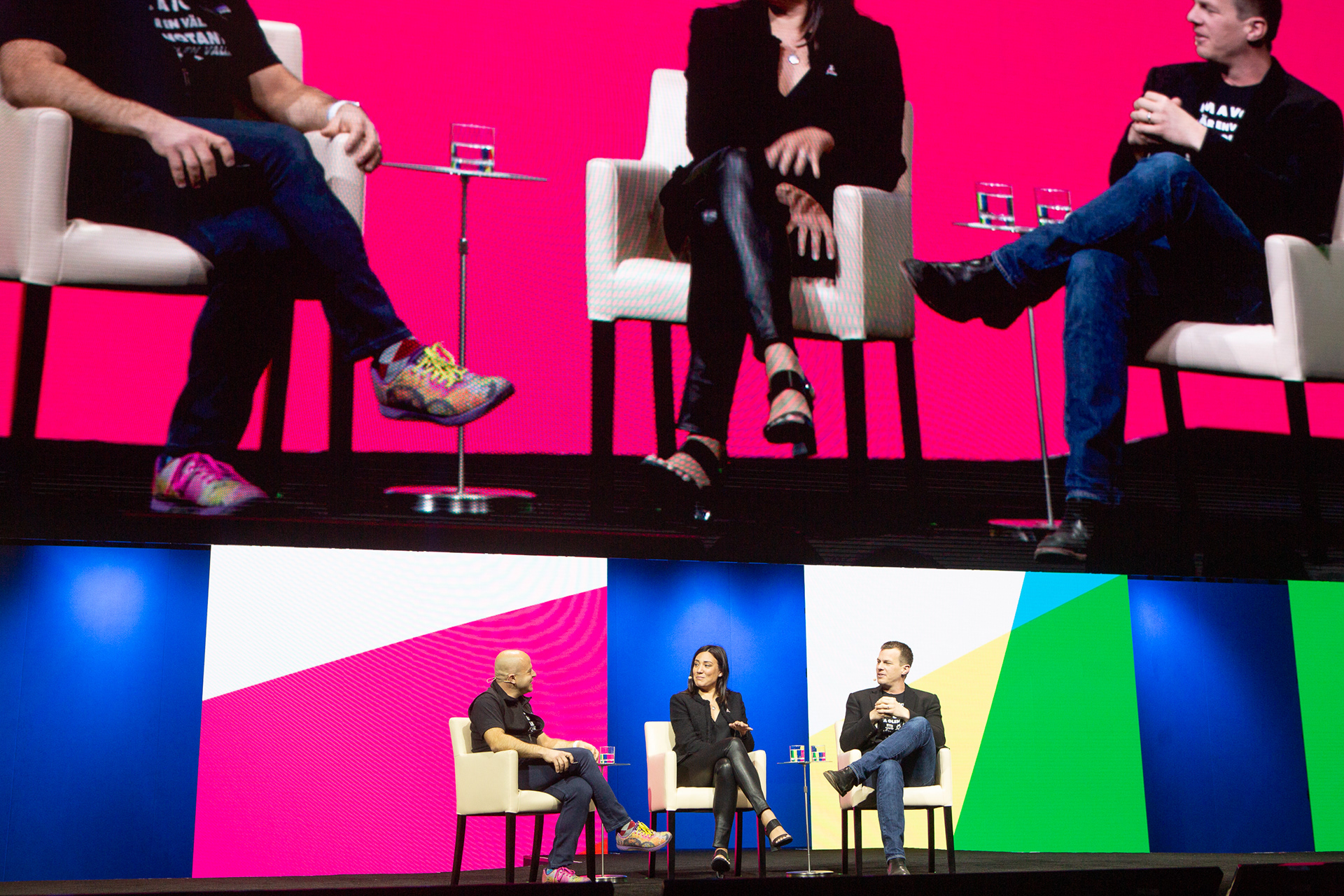 Three-person panel on stage with colorful geometric backdrop and IMAG screen above